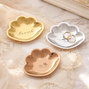 Può includere: Tre piattini portagioie a forma di conchiglia con finiture oro, argento e oro rosa. Il piattino dorato è inciso con il nome Hannah, quello argentato riporta il testo m.r. 01.18.2022 con due anelli e quello in oro rosa mostra S+R 11.06.2024.