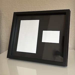 May include: A black picture frame with two white mat openings. The frame is rectangular and angled, with a larger, vertical opening and a smaller, horizontal opening. The frame is on a white surface.