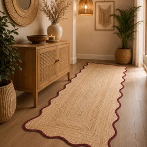 May include: A rectangular woven jute runner rug featuring a natural tan fiber texture and a scalloped edge finished with a dark burgundy border. The rug lies flat on a wooden floor, showcasing a consistent weave pattern and a decorative wavy trim.