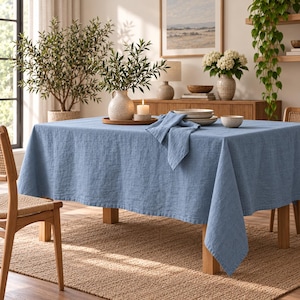 May include: A rectangular blue linen tablecloth draped over a wooden table. The fabric has a visible woven texture and a matte finish. A matching blue linen napkin is folded and placed on top of the tablecloth near the center of the table.