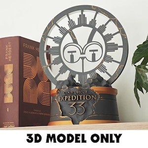 May include: A decorative, dark gray and gold-colored 3D model featuring a circular design with a central emblem and the text "CLAIR OBSCUR EXPEDITION 33". Two "Frank Herbert" books are visible.