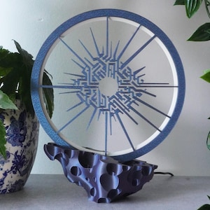 Könnte beinhalten: Eine moderne, kreisförmige Lampe mit blau-weißem Design, die auf einem dunkelvioletten Sockel steht. Die Lampe hat ein geometrisches Muster und wird mit einer Topfpflanze und hängendem Grün gezeigt.