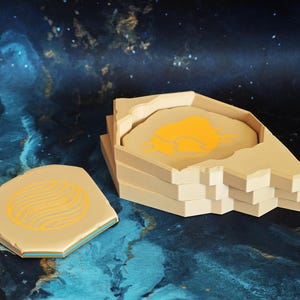 May include: A set of beige coasters with a geometric design in yellow. One coaster is separate, while the others are stacked in a matching holder. The coasters have a blue edge and are set against a dark blue and gold background.