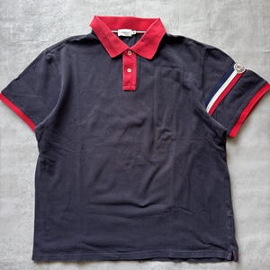 May include: A dark navy blue short-sleeved polo shirt featuring a bright red collar and a matching red button placket with two buttons. The left sleeve displays a red, white, and blue striped band with a circular logo patch near the edge.