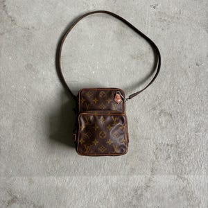 May include: A brown Louis Vuitton monogram canvas crossbody bag featuring a rectangular main compartment, a smaller front pocket, brown leather trim, and a thin adjustable shoulder strap with a metal buckle and hardware.