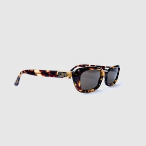 May include: Tortoiseshell sunglasses with dark tinted lenses and a thick, rounded frame. The temples feature a gold-tone metal accent with a prominent G logo integrated into the hinge area, connecting the frame front to the ear pieces.
