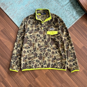 Peut inclure: Un pull en polaire camouflage avec un col boutonné et une petite poche. Le pull présente un motif de formes marron, vertes et beiges. Le col, la poche et la bordure sont d'un jaune néon vif.