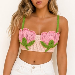 May include: A cropped crochet top featuring a light pink base with two large, three-dimensional pink tulip motifs on the front. The tulips have green leaves and stems, and the top is held up by two green crochet shoulder straps.