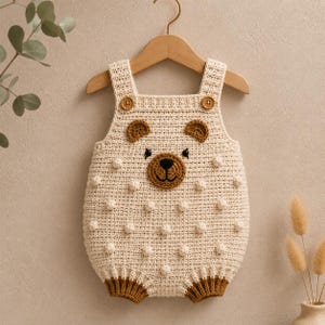 May include: A cream-colored crochet romper featuring a bear face design on the front. The garment has two brown wooden buttons at the shoulder straps, a textured bobble stitch pattern, and brown ribbed leg cuffs. The bear face includes brown ears.