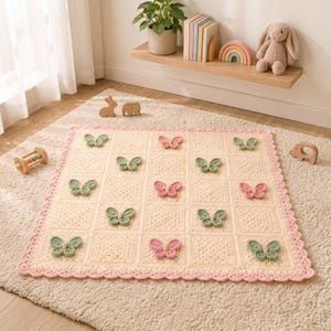 Puede incluir: Una manta cuadrada de ganchillo en hilo color crema con una cuadr&iacute;cula de cuadrados texturizados. La manta est&aacute; decorada con aplicaciones de mariposas tridimensionales en verde salvia y rosa suave, con un borde festoneado en rosa.