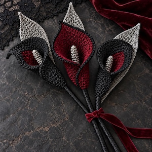 May include: Three handcrafted crochet calla lilies featuring black and deep red petals with silver-toned centers and leaves. The stems are wrapped in black material and tied together with a dark red velvet ribbon, creating a floral arrangement.