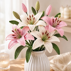 May include: A bouquet of crocheted lily flowers in shades of white and pink with dark brown stamens and green leaves. The flowers are arranged in a white ceramic vase with a ribbed texture, featuring several closed buds and fully bloomed petals.