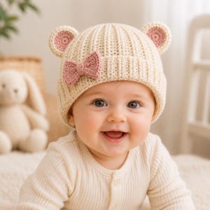 May include: A cream-colored crocheted baby hat featuring two small rounded ears on top with pink centers and a matching pink crocheted bow attached to the side. The hat has a ribbed texture and is worn by a smiling infant in a cream-colored shirt.
