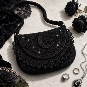 以下が含まれることがあります： フラップに三日月と星の模様があしらわれた黒いかぎ針編みのショルダーバッグ。バッグの底辺にはスカラップ状のレース風の縁取りがあり、太いかぎ針編みのストラップが付いています。小さな銀色の星のスタッドがデザインのアクセントになっています。