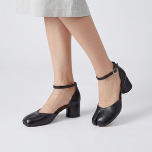May include: Black leather heeled sandals with a unique split-toe design. The sandals feature a thick, block heel and an ankle strap with a buckle closure. The shoes are a solid black color.