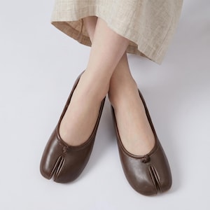 Könnte beinhalten: Ein Paar dunkelbraune Tabi-Ballerinas aus Leder mit markantem Zehenschlitz-Design, glatter Oberfläche und einem kleinen dekorativen Knoten-Detail in der Mitte des Vorderblatts. Diese Slipper haben eine flache Silhouette und eine runde Öffnung.
