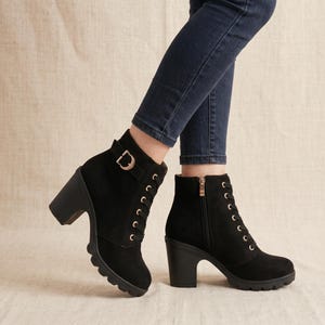 Könnte beinhalten: Ein Paar schwarze Stiefeletten mit hohem Blockabsatz, profilierter Sohle und seitlichem Schnallenriemen. Die Stiefel haben eine Schnürung vorne mit goldfarbenen Ösen und einen seitlichen Reißverschluss, getragen zu einer dunkelblauen Jeans.
