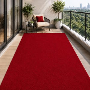 May include: A rectangular area rug in a solid, vibrant red color. The rug features a low-pile texture and is positioned flat on a light-colored wooden floor surface, extending from the foreground toward the middle of the outdoor balcony space.
