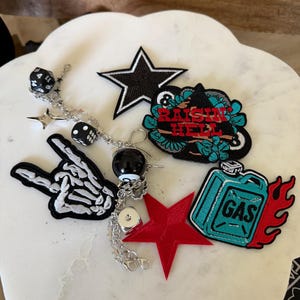 Op de afbeelding: Een verzameling geborduurde patches en een zilverkleurige bedelketting. De patches bevatten een zwarte ster, een skeletthand, een rode ster, een blauwgroene jerrycan met rode vlammen en een zwarte patch met de tekst RAISIN HELL. De ketting heeft dobbelsteenbedels.