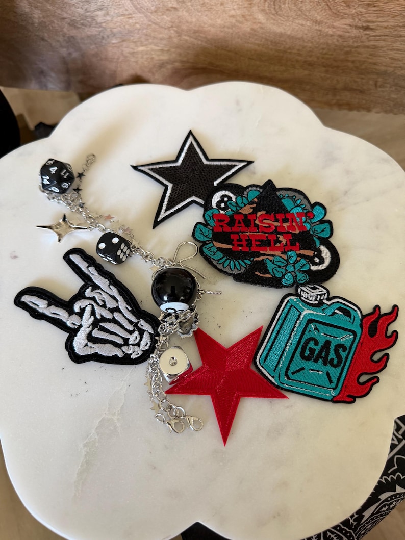 Op de afbeelding: Een verzameling geborduurde patches en een zilverkleurige bedelketting. De patches bevatten een zwarte ster, een skeletthand, een rode ster, een blauwgroene jerrycan met rode vlammen en een zwarte patch met de tekst RAISIN HELL. De ketting heeft dobbelsteenbedels.