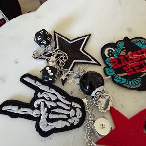 Op de afbeelding: Een verzameling decoratieve patches en bedels, waaronder een skeletthand, een zwarte ster, een rode ster en een ronde patch met de tekst "RAISE HELL". De items zijn verbonden door zilveren kettingen met dobbelstenen, een achtbal en kleine steraccenten.