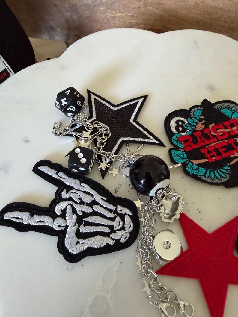Op de afbeelding: Een verzameling decoratieve patches en bedels, waaronder een skeletthand, een zwarte ster, een rode ster en een ronde patch met de tekst "RAISE HELL". De items zijn verbonden door zilveren kettingen met dobbelstenen, een achtbal en kleine steraccenten.