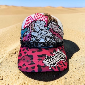 May include: A white trucker hat featuring a pink leopard print brim, a black chain detail, and various patches including a rider on an ATV, checkered flags, and the text Born To Race and Checkered Inbothered with a tire tread graphic.