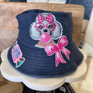 May include: A dark blue denim bucket hat featuring an embroidered patch of a grey poodle wearing pink heart-shaped glasses and a matching bow, blowing a pink bubble. A large pink bow patch and a smaller rectangular bubble gum patch are attached.