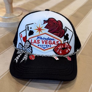 May include: Black and white trucker hat with a mesh back. The front panel features a Las Vegas design with playing cards, a rose, and the text "Fabulous Las Vegas Masters". A chain with charms adorns the brim.