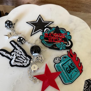 May include: A collection of embroidered patches and a silver-toned charm bracelet. The patches include a black star, a teal gas can with red flames and the text "GAS", and a black shape with teal flowers and the red text "RAISIN HELL". A skeleton hand patch and a red star patch are also included.