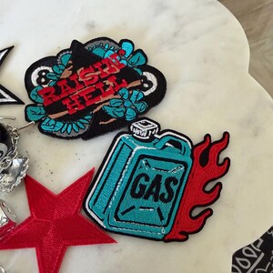 Op de afbeelding: Een verzameling geborduurde patches, waaronder een zwarte patch met de rode tekst RAISIN HELL omringd door turquoise bloemen, een turquoise jerrycan-patch met rode vlammen, een rode vijfpuntige ster en een gedeeltelijk zichtbare zwart-witte patch.