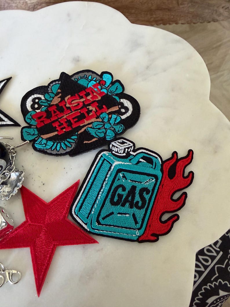 Op de afbeelding: Een verzameling geborduurde patches, waaronder een zwarte patch met de rode tekst RAISIN HELL omringd door turquoise bloemen, een turquoise jerrycan-patch met rode vlammen, een rode vijfpuntige ster en een gedeeltelijk zichtbare zwart-witte patch.
