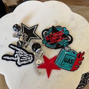 Op de afbeelding: Een verzameling geborduurde patches en bedels, waaronder een skeletthand, een zwarte ster, een rode ster, een blauwgroene jerrycan met vlammen en een patch met de tekst "RAISE HELL". Zilveren kettingen verbinden bedels in de vorm van dobbelstenen.
