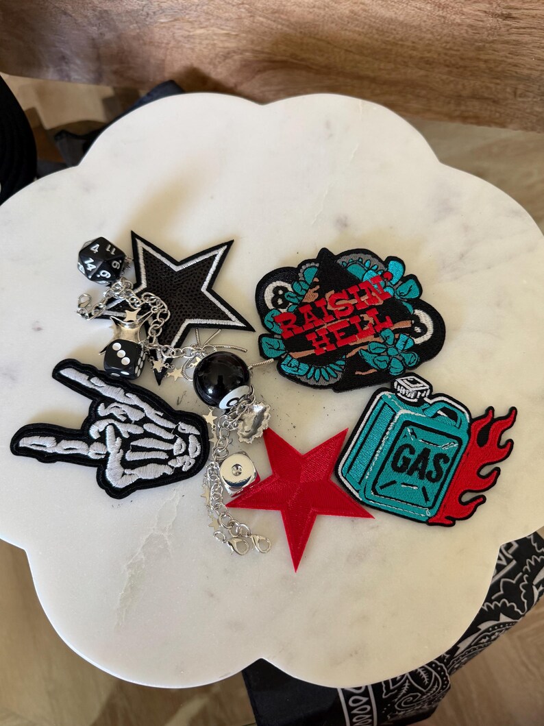 Op de afbeelding: Een verzameling geborduurde patches en bedels, waaronder een skeletthand, een zwarte ster, een rode ster, een blauwgroene jerrycan met vlammen en een patch met de tekst "RAISE HELL". Zilveren kettingen verbinden bedels in de vorm van dobbelstenen.