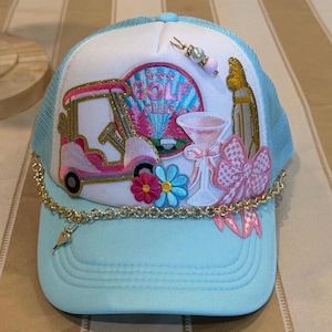Peut inclure: Casquette de camionneur bleu clair et blanche ornée sur le thème du golf. Elle présente une voiturette de golf rose, un verre à martini et un nœud rose. Une chaîne et un charme dorés pendent de la visière. Le chapeau a un dos en filet.