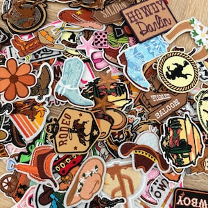 May include: A collection of embroidered patches featuring western themes, including cowboy boots in pink and blue, cowboy hats, cacti, horses, stars, and signs with text such as Howdy Darlin, Rodeo, and Saloon, all arranged in a dense, overlapping pile.