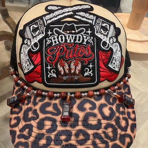May include: A beige and black trucker hat with a leopard print brim. The hat features a patch with crossed pistols, the words "HOWDY P*TOS", and cowboy boots. A beaded chain with miniature liquor bottle charms adorns the brim.