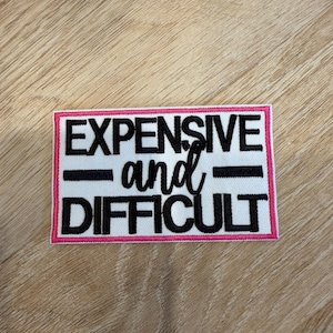 Peut inclure: Un écusson rectangulaire brodé avec un fond blanc et une bordure rose. Le texte indique EXPENSIVE en lettres capitales noires, suivi du mot and en écriture cursive noire, et du mot DIFFICULT en lettres capitales noires en dessous.