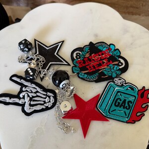 Op de afbeelding: Een verzameling geborduurde patches en een zilverkleurige kettinghanger met een zwarte ster, een skeletthand, een rode ster, een jerrycan met vlammen en een patch met de tekst RAISE HELL, samen met dobbelstenen en een acht-ball bedel.