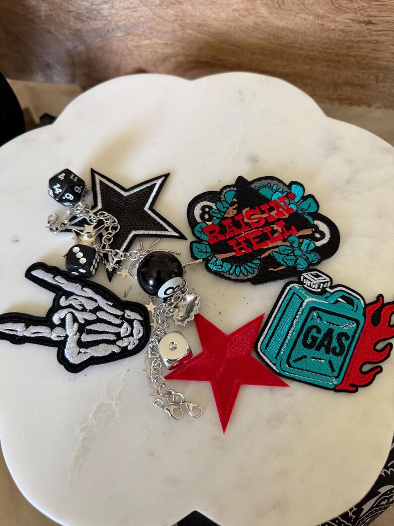 Op de afbeelding: Een verzameling geborduurde patches en een zilverkleurige kettinghanger met een zwarte ster, een skeletthand, een rode ster, een jerrycan met vlammen en een patch met de tekst RAISE HELL, samen met dobbelstenen en een acht-ball bedel.