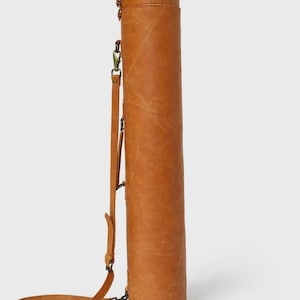 May include: A cylindrical, tan leather carrying case with a shoulder strap. The case has a top flap with a buckle closure and a long, adjustable strap. The leather has visible grain and a natural finish. The case is designed to hold long, slender items.