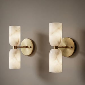 May include: Two wall sconces featuring a circular brushed brass backplate and a horizontal arm supporting two vertically stacked, cylindrical, translucent white stone shades with grey veining. The light fixtures emit a soft, warm glow.