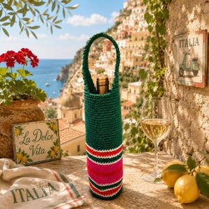 May include: A dark green crochet bottle holder with a handle, featuring horizontal stripes in white, red, and bright pink at the base. The holder contains a glass bottle with a gold-colored cap, standing upright on a textured stone surface.