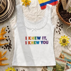May include: A white ribbed tank top with lace trim along the scoop neckline and a small center bow. The text I KNEW IT, I KNEW YOU is embroidered on the chest in multi-colored rainbow lettering. The garment features a sleeveless design.