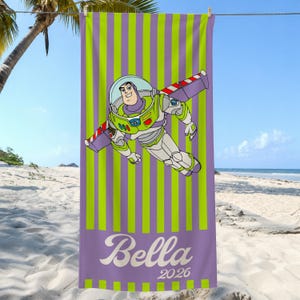 May include: A rectangular beach towel featuring a vertical lime green and purple striped pattern. The center displays a graphic of a space ranger character in a white, green, and purple suit. The name Bella and the year 2026 are printed in white.