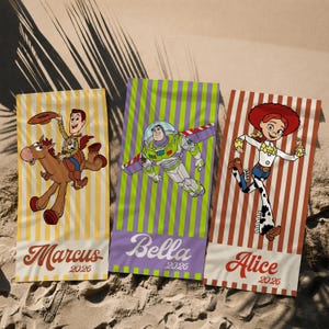 May include: Three rectangular beach towels featuring Toy Story characters. The left towel has yellow stripes, Woody on Bullseye, and the name Marcus 2026. The center towel has green stripes, Buzz Lightyear, and Bella 2026. The right towel has red stripes, Jessie, and Alice 2026.
