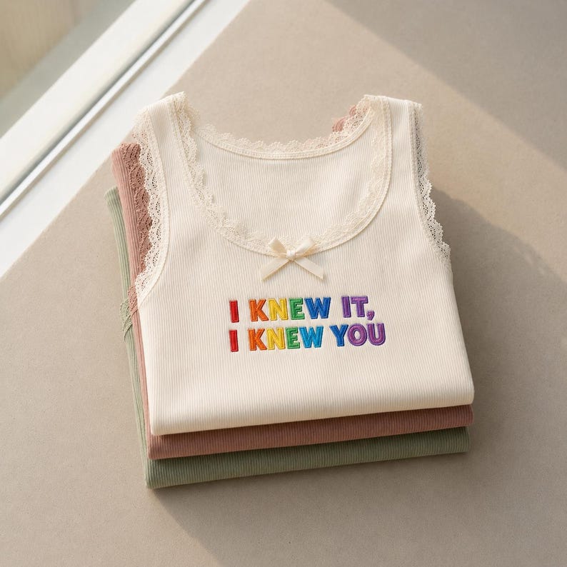 May include: A stack of three ribbed tank tops featuring lace trim along the scoop neckline and a small center bow. The top cream-colored tank displays the embroidered text I KNEW IT, I KNEW YOU in multi-colored rainbow lettering.