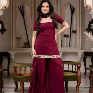 May include: A deep maroon kurta set featuring a square neckline, short sleeves, and wide-leg palazzo pants. The garment is accented with a cream-colored decorative trim along the neckline, sleeve cuffs, and hemline of the tunic.