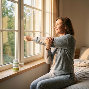 May include: A woman in gray activewear stretches near a window. A white bottle with a green and yellow label, and a glass of water sit on the windowsill. The window overlooks a green, leafy outdoor scene.