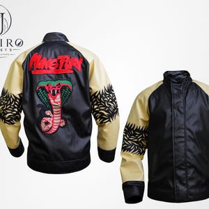 Può includere: Giacca nera e beige con un disegno di cobra sul retro. La giacca ha un collo alto e finiture nere. Le maniche sono beige con motivi neri a forma di foglia. La parola "MangTien" è ricamata in rosso.